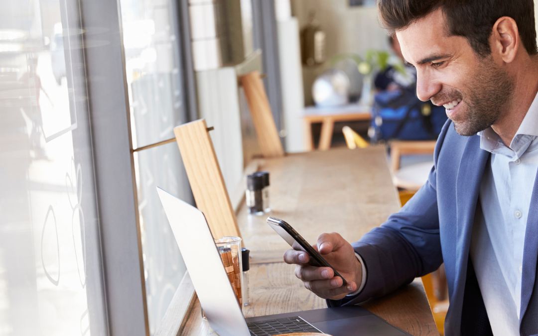 Profissional sorrindo ao usar o celular em um café, com notebook na mesa